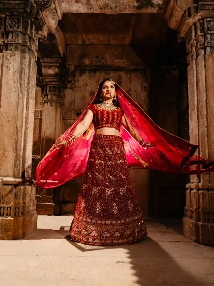 A full-length portrait of the bride, showcasing the magnificent detail of her red lehenga against the ancient stone. The flowing dupatta adds a touch of grace and movement to this regal shot.