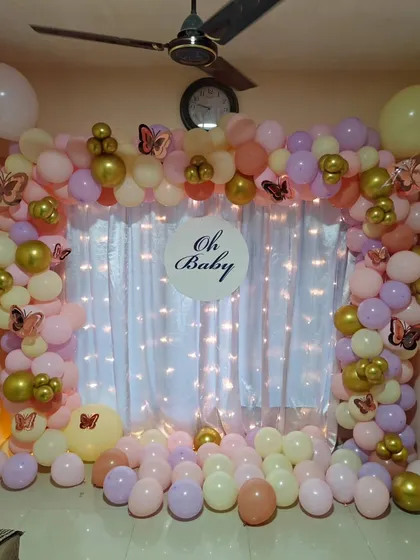 An 'Oh Baby' photo corner created for a home baby shower. It features a full balloon wall in pastel shades with gold accents, butterfly stickers, and a fairy light curtain.