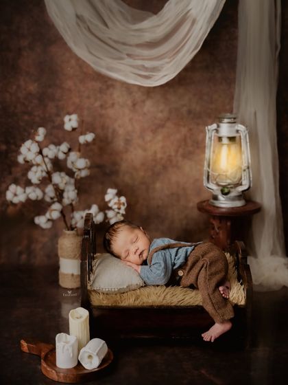 A rustic and cozy newborn setup featuring a miniature wooden bed. The baby sleeps soundly, dressed in tiny overalls, with a soft lantern glow adding to the warm, peaceful ambiance.
