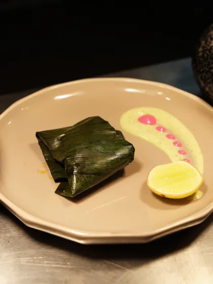 A fish dish wrapped in a banana leaf, presented with a colourful sauce and a slice of lemon. The plating is clean, modern, and appetizing.