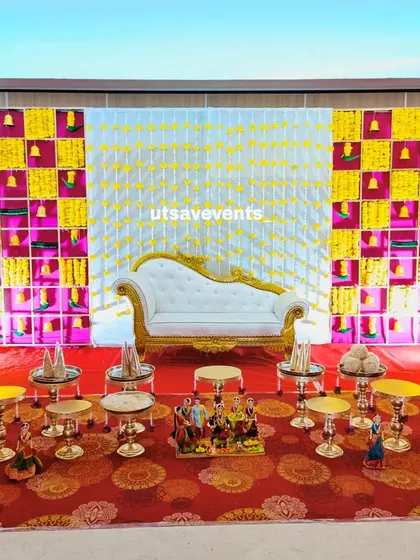 A beautiful baby shower stage with a checkered floral backdrop and a white divan. The floor is decorated with traditional dolls and offerings on small brass tables.