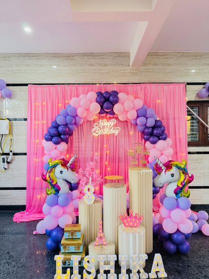A clear, well-lit photo of the unicorn theme birthday decoration. The pink and purple balloon arch is perfectly formed around the pink curtain backdrop with fairy lights.