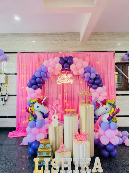 A clear, well-lit photo of the unicorn theme birthday decoration. The pink and purple balloon arch is perfectly formed around the pink curtain backdrop with fairy lights.