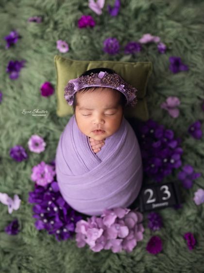 A sweet newborn portrait with a date block marking the special day. The baby is wrapped in purple and surrounded by matching flowers, perfect for remembering every detail.