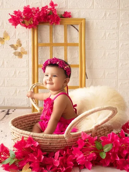 A happy baby girl in a bright pink outfit sits in a basket surrounded by bougainvillea flowers. A vibrant and cheerful half-birthday portrait.