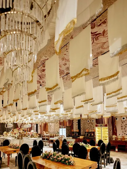An elegant indoor dining setup with a ceiling of hanging white and gold fabric panels and stunning crystal chandeliers.