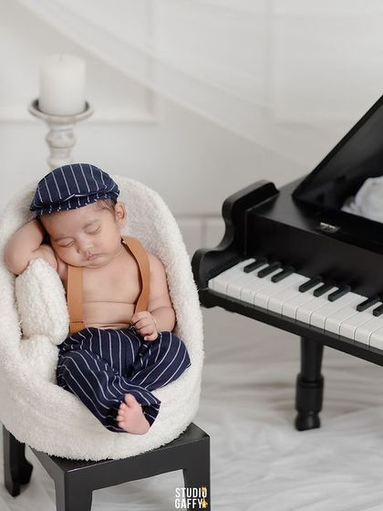 A little musician in the making, this newborn is styled with a tiny cap and suspenders, sleeping peacefully in a chair next to a mini piano.