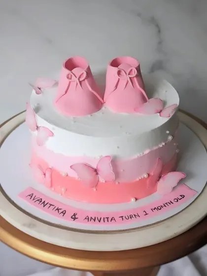 Celebrating one month of twin girls with this sweet pink cake. The design features two pairs of baby booties and a cascade of pink butterflies.