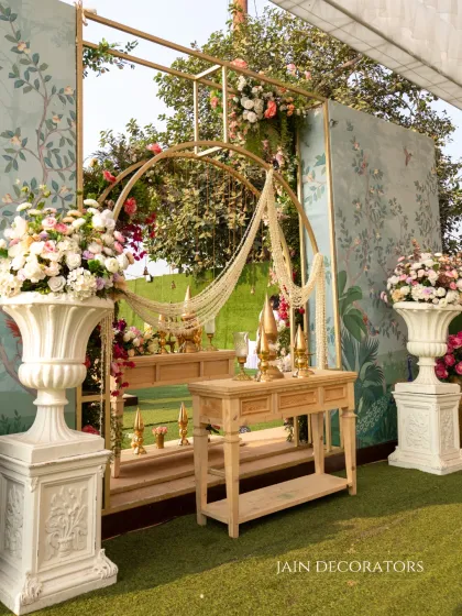 A beautiful and artistic photo corner. A golden circular frame is draped with pearls and set against a hand-painted backdrop, flanked by classic floral urns.