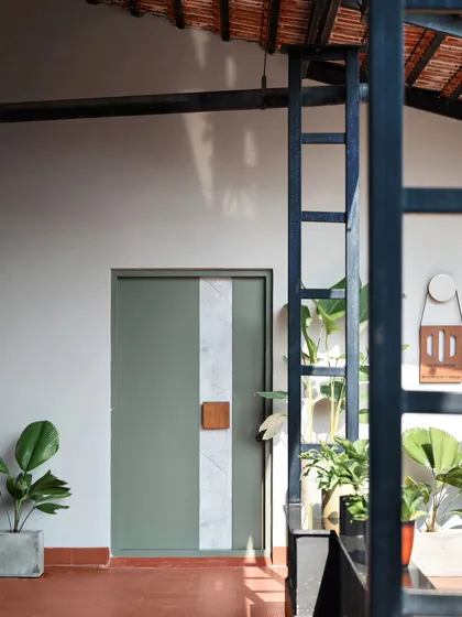 This perspective highlights the interplay of materials at the office entrance. The dark metal frame of the structure contrasts with the light walls and green door, while the natural light casts soft shadows, adding depth and character to the space.