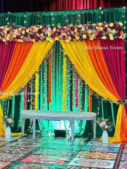 A grand stage for a Mehendi or Sangeet function. The backdrop features layers of colorful drapes in green, orange, and pink, with a floral border on top and a beautiful printed carpet.