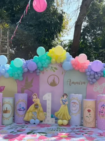 An outdoor Disney Princess-themed first birthday party, featuring characters like Belle and Snow White on custom pedestals and a pastel balloon garland.