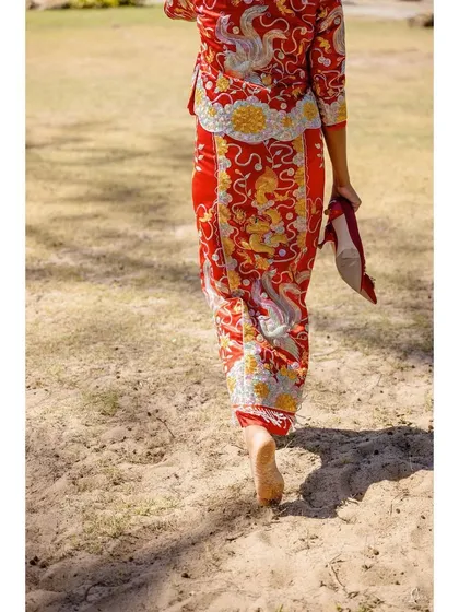 The bride, Shachi, walks barefoot on the sand in Phuket, holding her shoes. This simple, candid moment captures the relaxed and beautiful atmosphere of a beach wedding.