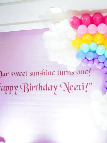 A close-up of the "Our sweet sunshine turns one!" backdrop. The clear, elegant font against the pink background is framed by a vibrant rainbow balloon arch, creating a perfect photo moment.