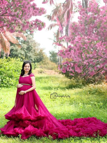A regal portrait of a mother-to-be in a wine-colored ruffled gown and crown, set in a beautiful pink-themed garden.