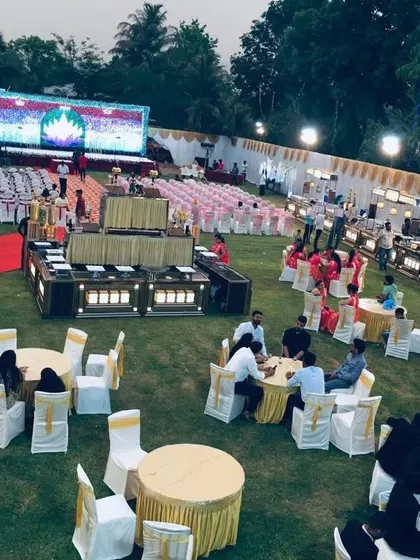 An aerial view of a large event at Shrusti Farmhouse & Resort in Panvel. You can see the spacious lawn, stage with a large LED screen, and separate seating and dining areas for a massive gathering.