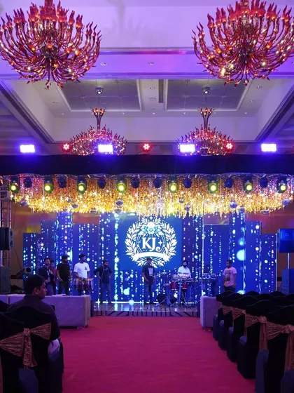 A grand Sangeet stage setup with a large LED screen for the couple's monogram, flanked by smaller screens and topped with a canopy of golden lights and red chandeliers.