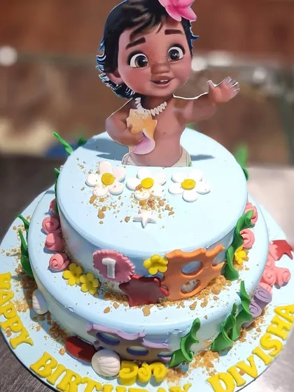 An adventurous Moana-themed cake for a first birthday. This two-tier cake is decorated with ocean-blue frosting, tropical flowers, and a baby Moana topper, ready to make a splash at the party.