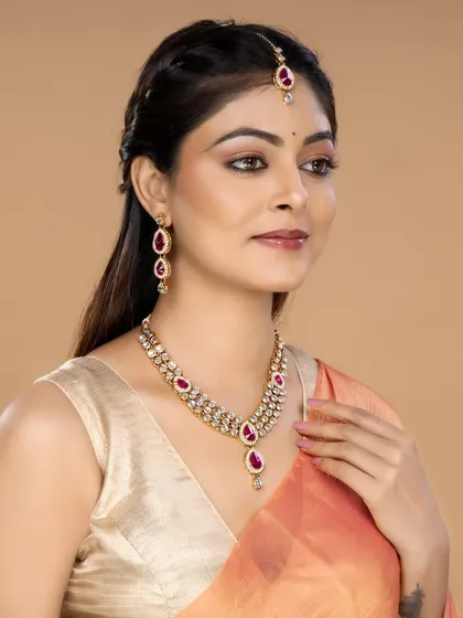 A model showcases a contemporary necklace set with pink gemstones and a matching maang tikka. The warm background and soft lighting complement the jewellery's tones, ideal for a festive collection.