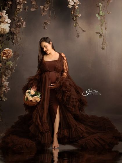 A solo portrait featuring a beautiful brown ruffled gown. The mom-to-be looks down lovingly at her bump, holding a basket of flowers, with a water reflection effect at her feet.