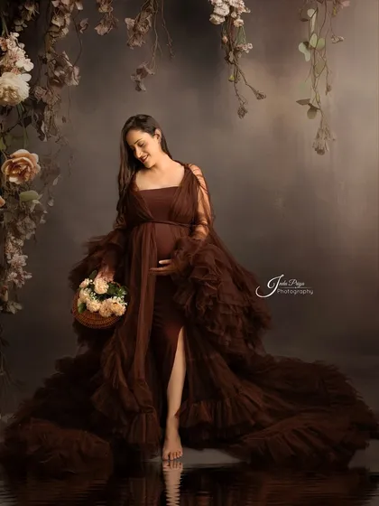 A solo portrait featuring a beautiful brown ruffled gown. The mom-to-be looks down lovingly at her bump, holding a basket of flowers, with a water reflection effect at her feet.