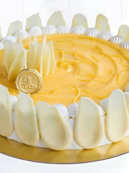 A beautiful cake showcasing a bright yellow curd topping, encircled by delicate white chocolate shards. Our iconic gold seal sits proudly in the center, a mark of our commitment to quality and elegance.