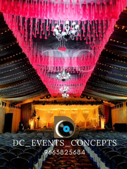 An elaborate indoor venue decoration featuring a stunning pink tassel chandelier, dark fabric ceiling drapes, and rows of string lights for a dramatic effect.