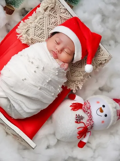 An overhead view of a swaddled newborn sleeping next to a friendly snowman prop in a bed of soft cotton snow.