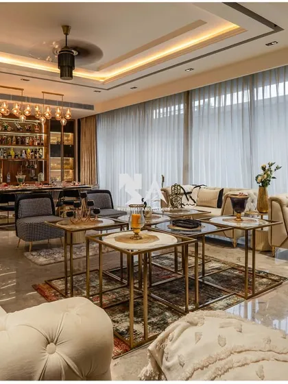 This elegant living area is designed for sophisticated entertaining, with a fully stocked bar and dining setup in the background. The tufted sofas, nested coffee tables, and sheer curtains create a layered look of luxury, with a view of the poolside just outside.