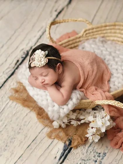 A newborn girl sleeping on her side in a basket, wrapped in a soft peach-colored fabric. The rustic textures and floral accents create a warm, bohemian-inspired baby photo.