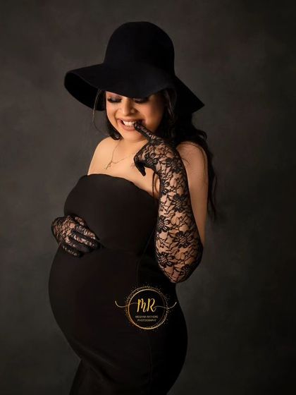 A close-up shot capturing a playful and happy moment. The mother-to-be smiles brightly, dressed in a stylish black outfit with a hat and lace gloves.