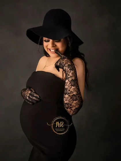 A close-up shot capturing a playful and happy moment. The mother-to-be smiles brightly, dressed in a stylish black outfit with a hat and lace gloves.
