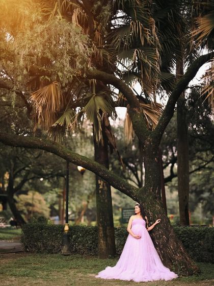 Captured during an outdoor session, the mother-to-be looks serene in a lavender gown, standing under the canopy of a large, beautiful tree. The golden hour light adds a magical glow to the scene.