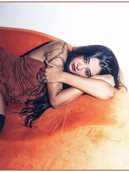 A more vulnerable and intimate pose from the same orange couch series. The model's curled-up position and soft expression create a different, more contemplative mood compared to the direct-gaze shots.