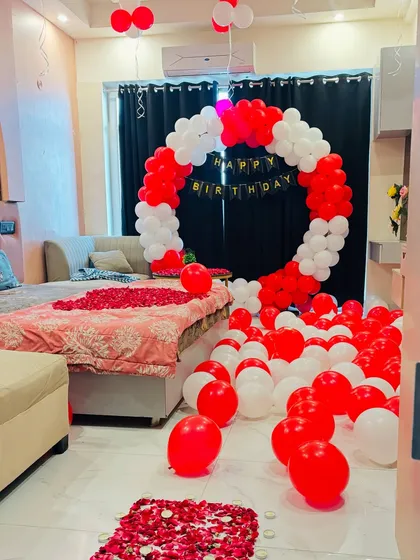 A birthday surprise in a bedroom with a circular balloon arch in red and white, and a floor covered in balloons and rose petals.