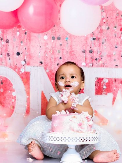 Getting hands-on with her first birthday cake. The sparkly pink backdrop and matching balloons make this a picture-perfect celebration for a little princess.
