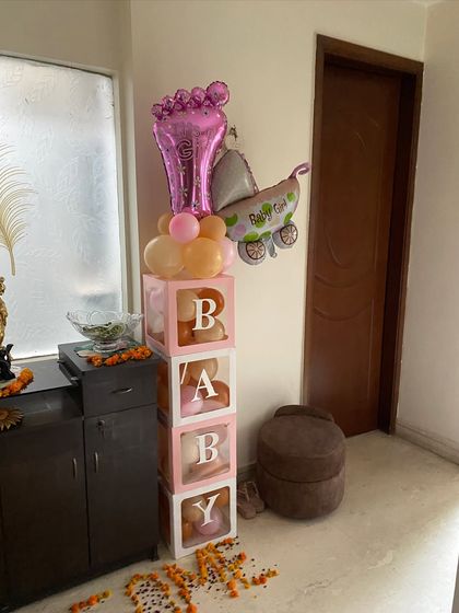 A corner decoration for a baby girl's arrival, featuring stackable 'BABY' boxes, and foil balloons shaped like a baby foot and a pram.
