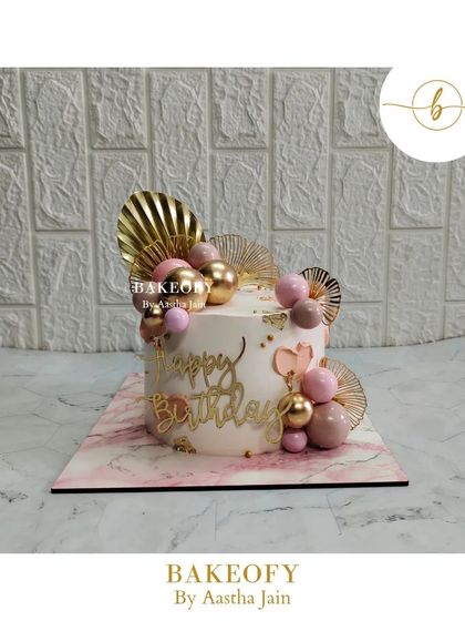 An aesthetically pleasing choco-vanilla cake. The design is simple but glamorous, with pink and gold spheres, golden fans, and a "Happy Birthday" topper.