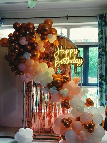 An elegant balloon arch setup in a living room with a view. This rose gold, peach, and white balloon arch is placed against a sheer curtain and a rose gold fringe backdrop.