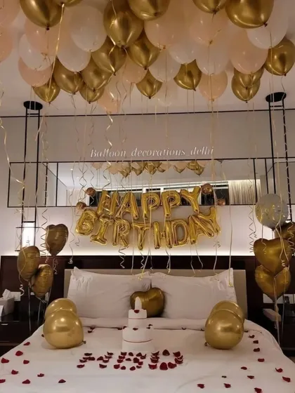A simple and elegant birthday room decoration with gold and white balloons, 'Happy Birthday' foil letters, and rose petals on the bed.