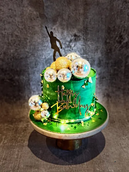 A vibrant disco-themed cake in sage green. The cake is decorated with gold and silver disco balls and a dancer silhouette topper, perfect for a retro-themed party.