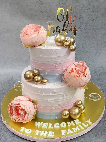A beautiful two-tiered "Welcome to the Family" cake. This tri-color icing design is adorned with large peonies and gold spheres, making it an elegant choice for a baby shower.