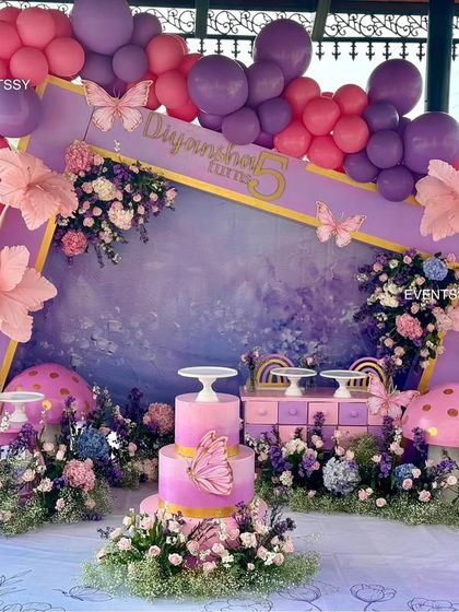A full view of the purple and pink floral party, showing the cake table and dessert stands integrated into the enchanting scene. The hand-painted backdrop adds an artistic, dreamy quality.