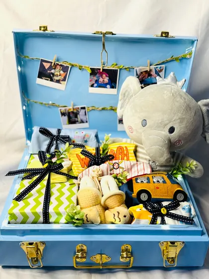 A gift hamper for a baby boy, packed in a beautiful blue trunk. It includes a soft elephant toy, a string of photos, and neatly wrapped clothing essentials.