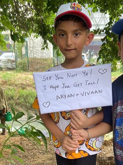 Two young boys hold a handmade sign that reads, "See you next yr! I hope you get tall," a heartfelt message to the sapling they just planted.