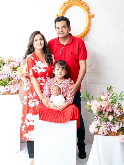 The complete family of four, beaming with joy. The older sister sits proudly with her new baby sibling, surrounded by her loving parents in a beautifully coordinated red-themed portrait.