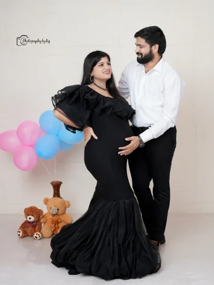 A loving couple's portrait in our studio, with the mother-to-be in a black mermaid-style gown, surrounded by gender-reveal balloons.