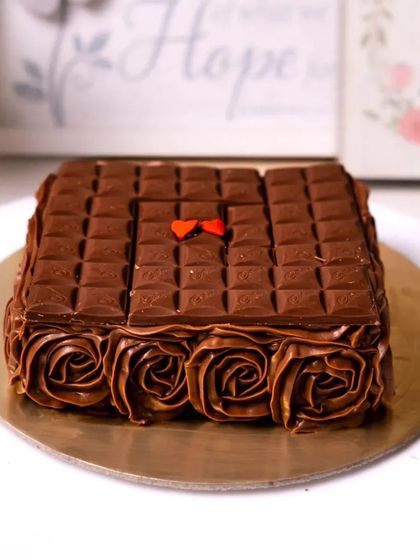 A square chocolate cake topped with chocolate bars and surrounded by piped chocolate rosettes.