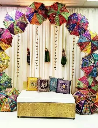 If you want a unique theme, I can create a beautiful Rajasthani setup. This backdrop features an arch made of colorful embroidered umbrellas and a comfortable seating area with traditional cushions.