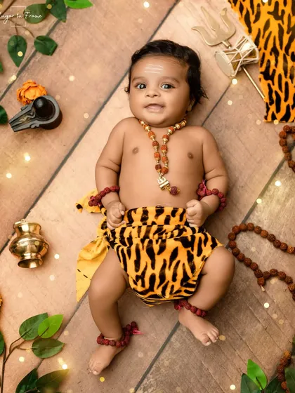 A happy little baby dressed as Lord Shiva, lying on a wooden floor setup with a shivling and other symbolic props.